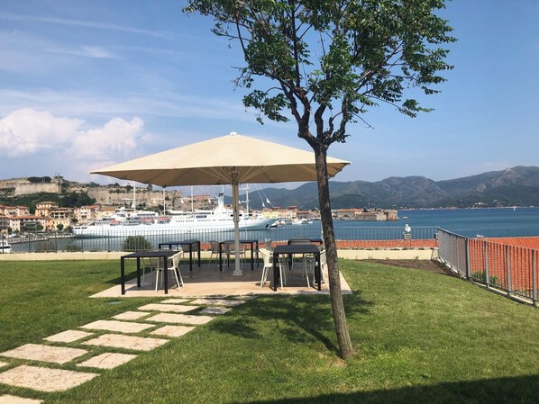 A patio with outdoor seating and a large umbrella, overlooking a marina with boats and a coastal town in the background.