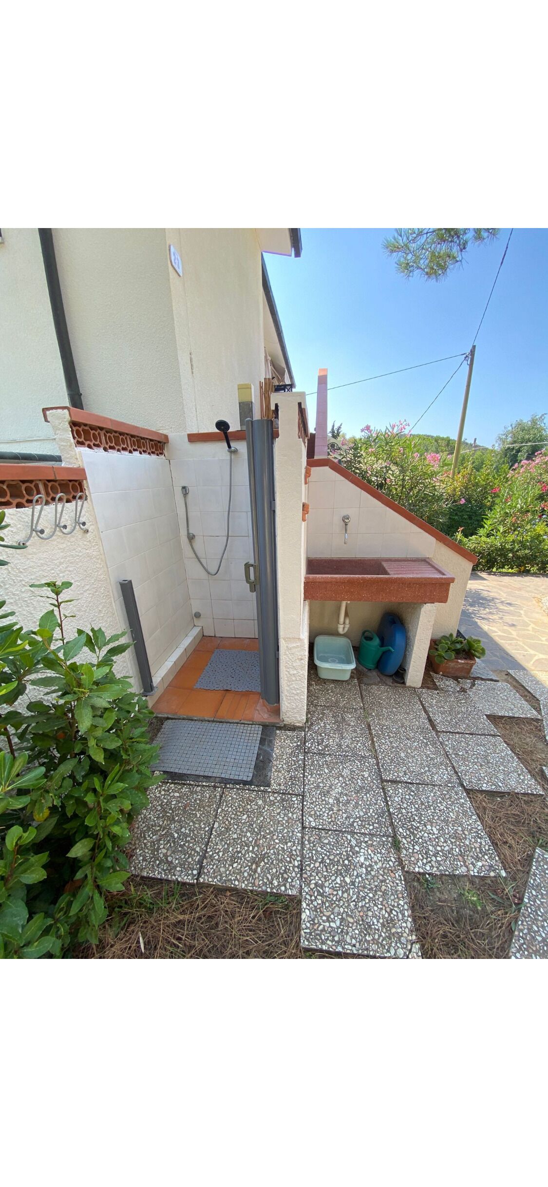 A small outdoor area with a shower, a sink, and a step leading to a door. There are plants and a tiled floor.