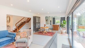 Un salon moderne avec un canapé bleu, un fauteuil gris, une table à manger en bois avec des chaises, un escalier et une vue sur le jardin grâce à de grandes fenêtres.