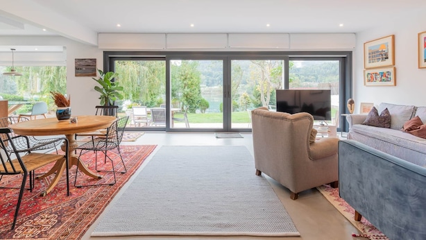 Un salon moderne doté d’une grande porte-fenêtre coulissante donnant sur un jardin, d’un canapé, d’un téléviseur et d’un coin repas avec des chaises et une table.