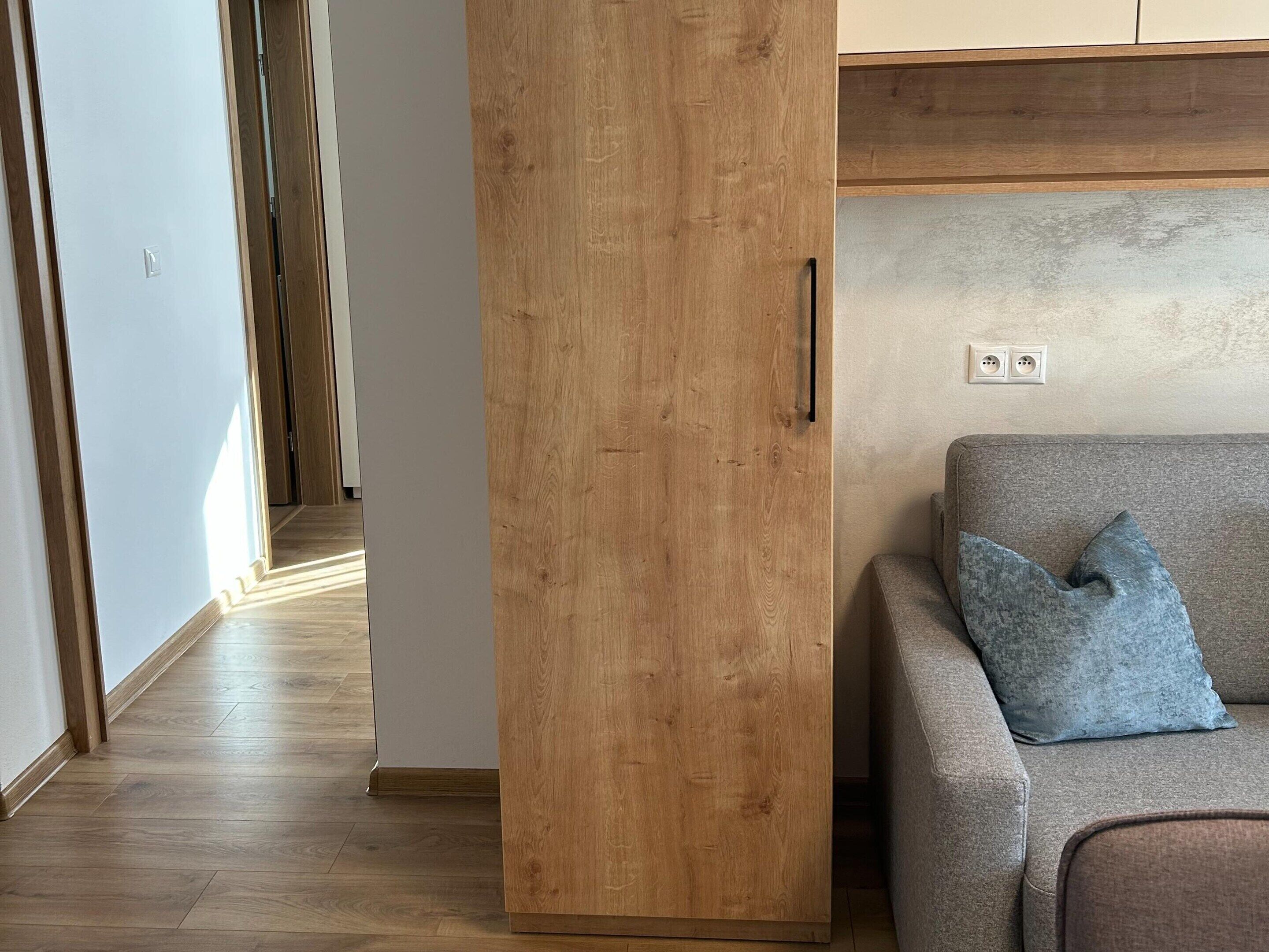 Una habitación con un armario de madera, un sofá gris con un cojín azul y un interruptor de luz en la pared.