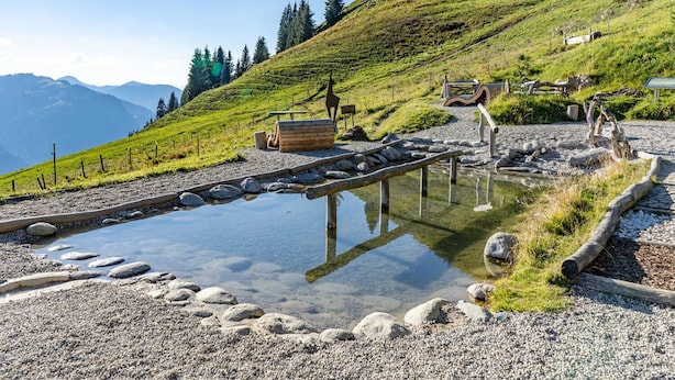 A clear outdoor pool with a wooden deck, surrounded by pebbles and a gravel path. - Apartment With ski Lift Right Outside the Door (Jochberg)