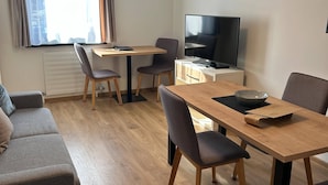 A modern living room with a sofa, a dining table with chairs, a TV, and a window with curtains. - Apartment With ski Lift Right Outside the Door (Jochberg)