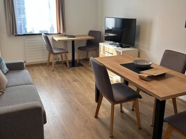 Ein modernes Wohnzimmer mit einer Couch, einem Esstisch mit Stühlen, einem Fernseher und einem Fenster mit Vorhängen.