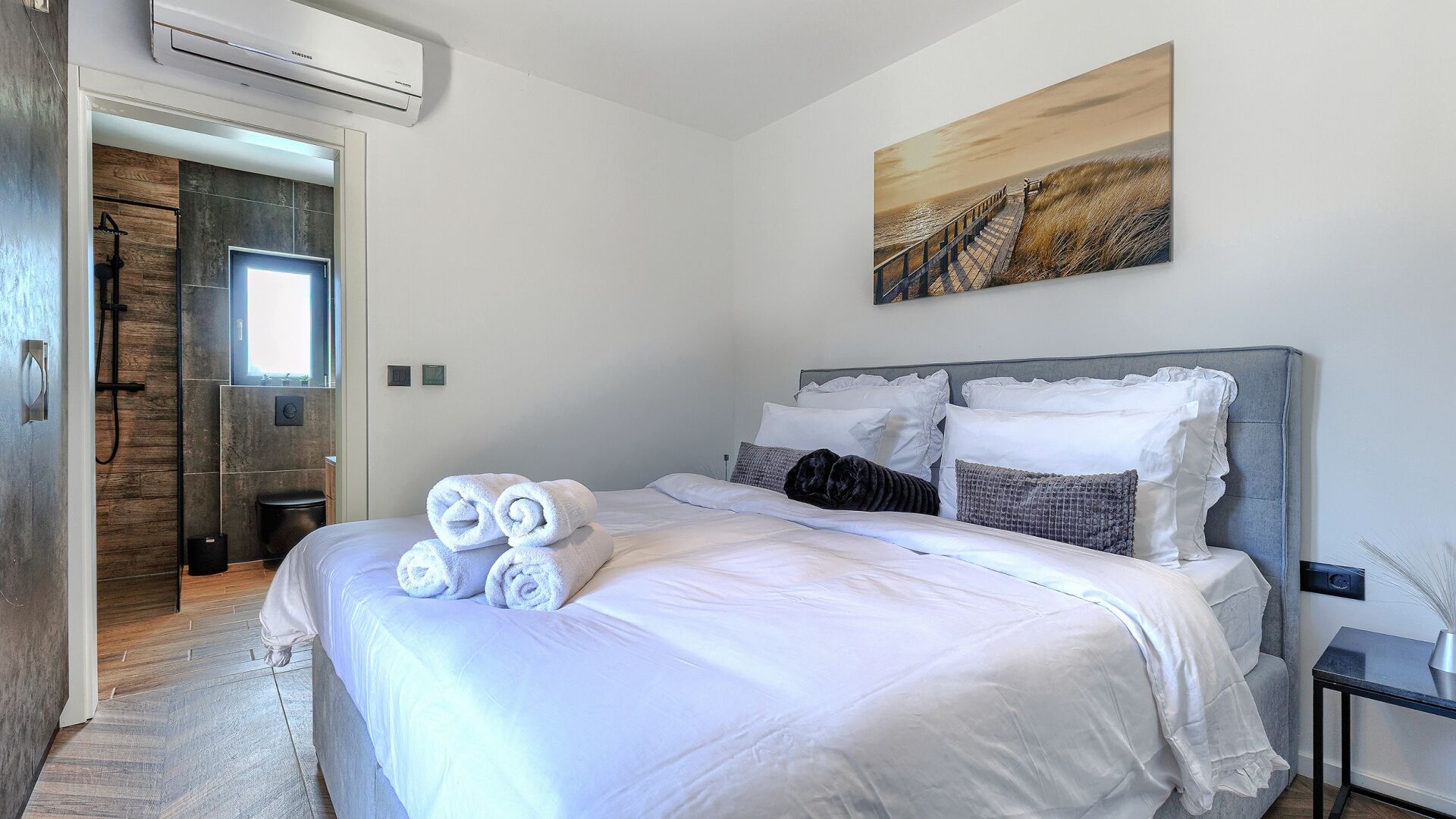 A modern hotel room with a large bed, a painting on the wall, and a view of the beach through the window.
