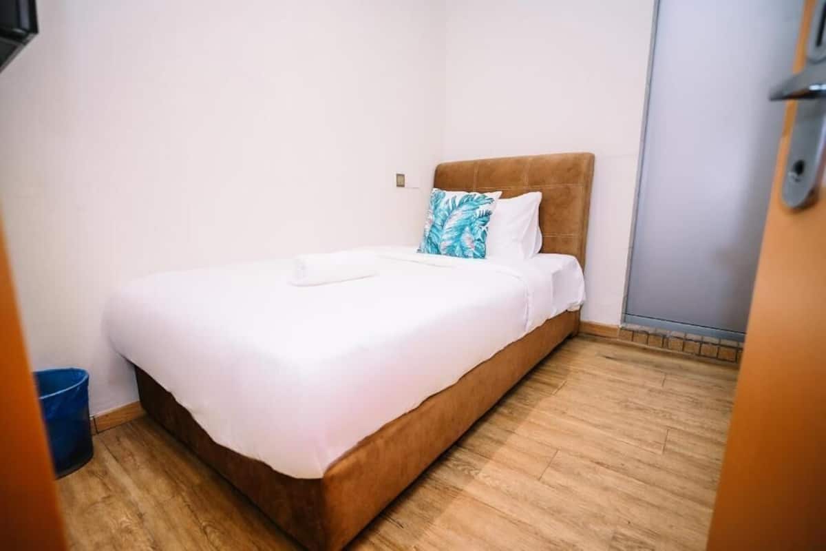 Una cama individual con cabecera de madera, ropa de cama blanca y un cubo de basura azul.