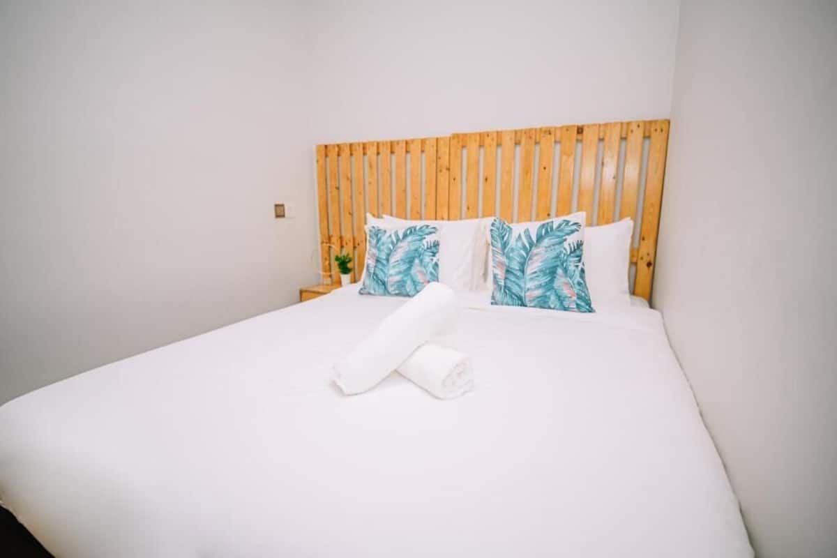 Una cama bien hecha con ropa de cama blanca y una toalla blanca enrollada, un cabecero de listones de madera y dos almohadas con un estampado de hojas tropicales.