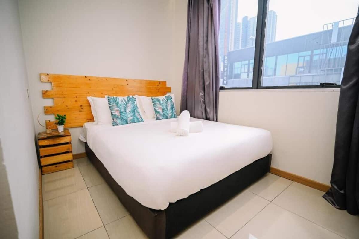 Habitación de hotel con cabecera de madera, una cama grande con ropa blanca, una mesita de noche con una planta y vista a un paisaje urbano a través de una ventana.