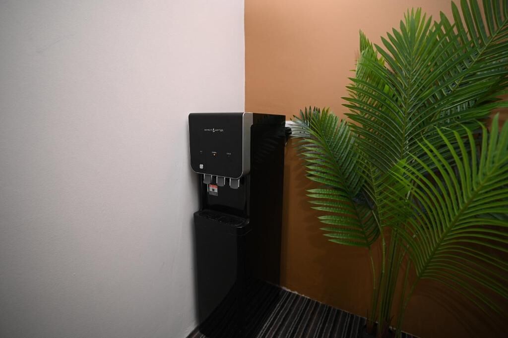 A self-service beverage dispenser mounted on a wall next to a trash bin and a potted plant.