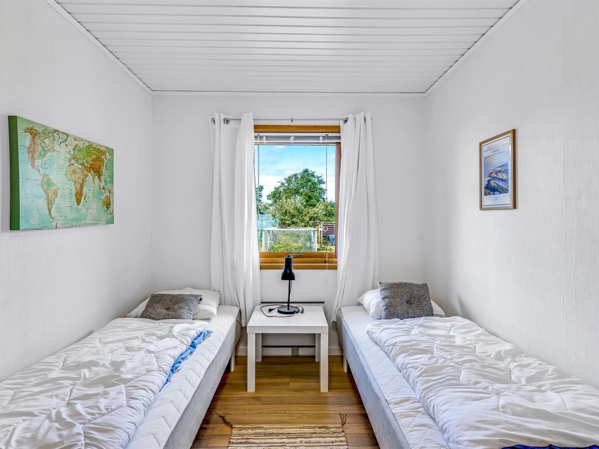 Ein Zimmer mit zwei Einzelbetten, einem kleinen Tisch mit einer Lampe, einer Weltkarte an der Wand und einem Fenster mit Blick auf Bäume und ein Gebäude.