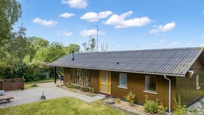 Yhden kerroksen korkuinen puutalo, jossa on kalteva katto, kuisti ja pieni puutarha, jossa kasvaa kukkia ja pensaita.
