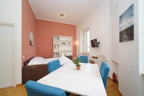 A room with a bed, a white table with blue chairs, a bookshelf, a TV, and a window.