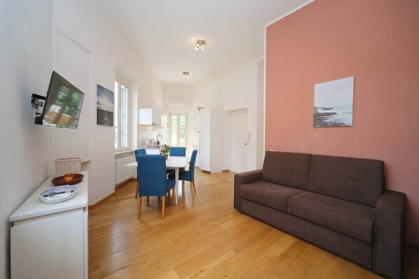 A modern living room with a brown sofa, a dining area with blue chairs, and a kitchen in the background.