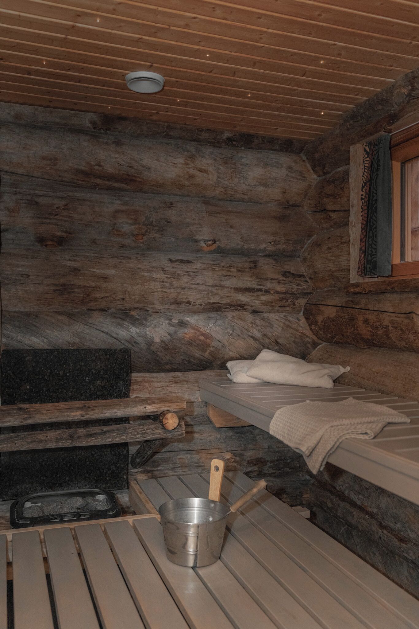 A log cabin interior with a wooden ceiling, a metal bucket, a bench, and a bed with a blanket.