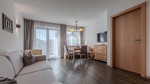 Ett modernt vardagsrum med en soffa, ett matbord och stolar. Det finns en trädörr, en tv som är monterad på väggen och ett fönster med gardiner.