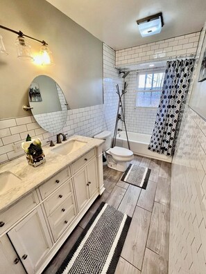 A bathroom with white subway tiles, a white sink with a mirror, a toilet, a shower with a patterned curtain, and a black and white striped rug.