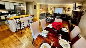 Uma cozinha moderna com uma ilha central, uma sala de jantar com uma mesa de madeira posta para uma refeição, e uma sala de estar com sofá e lareira.