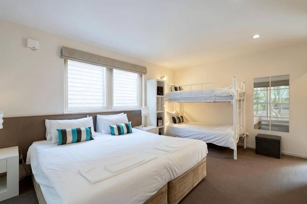 Habitación de hotel con una cama grande, una litera, una mesita de noche, una lámpara y una ventana con persianas.