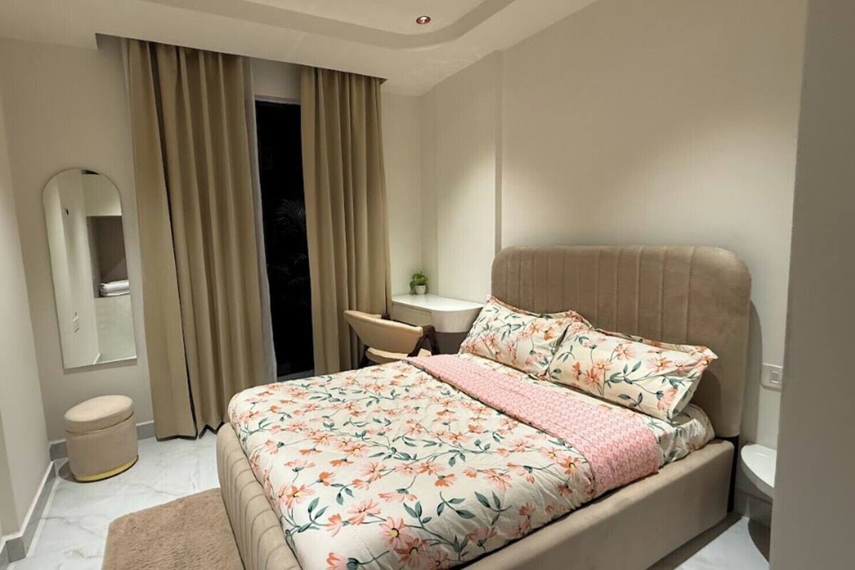 Un dormitorio con cama, mesita de noche, silla, espejo y ventana con cortinas.