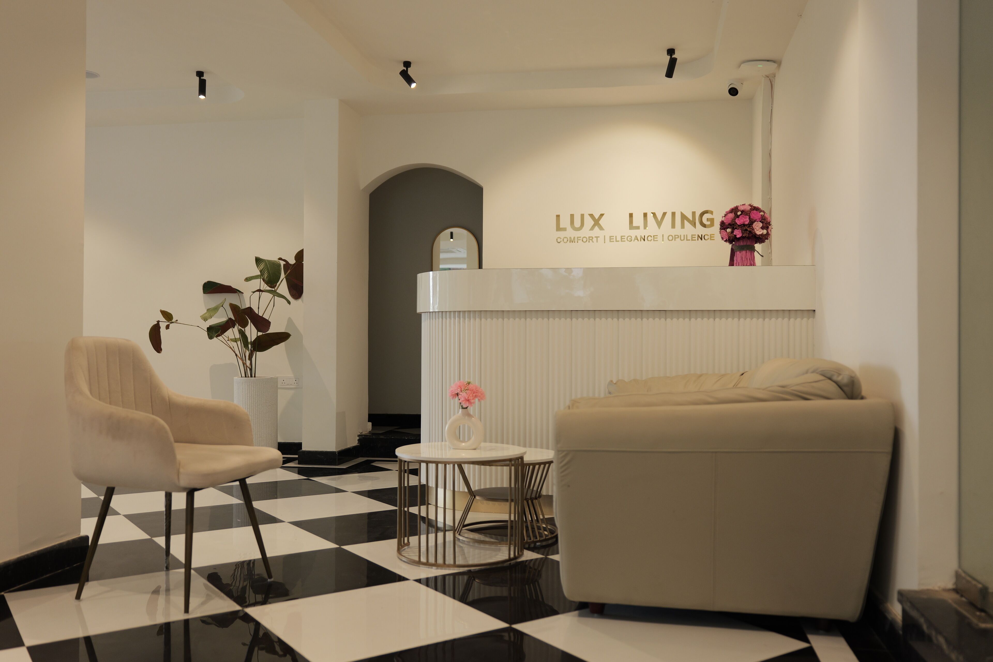 A modern lounge area with a checkered floor, a sofa, a chair, a small table with a vase of flowers, and a wall-mounted sign reading "LUX LIVING".