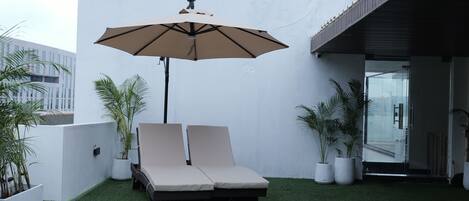 A rooftop lounge area with two lounge chairs, a large umbrella, and potted plants.