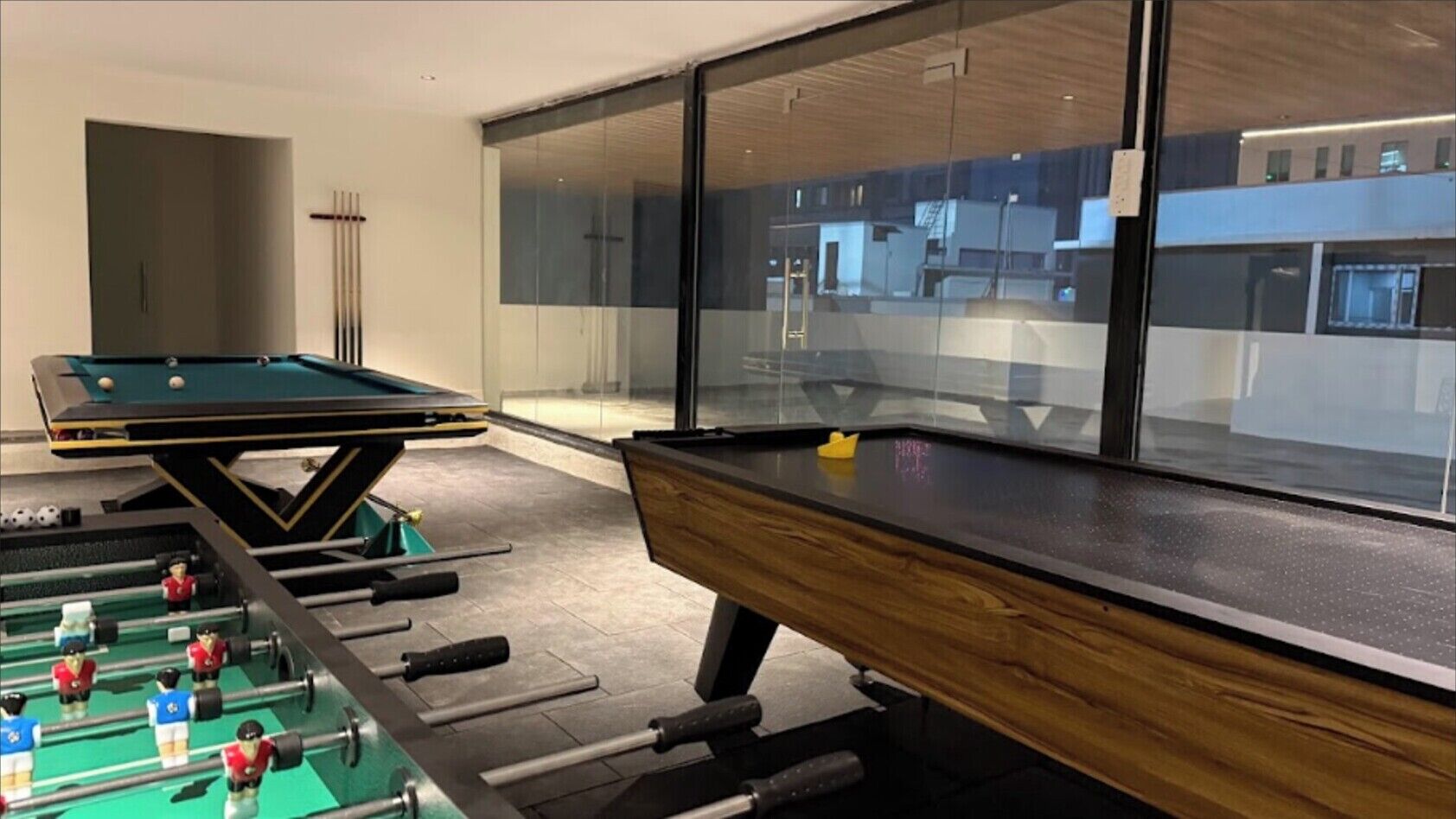 Een pooltafel met een tafelvoetbaltafel ernaast, een houten bankje, en uitzicht op het gebouw buiten door grote ramen.