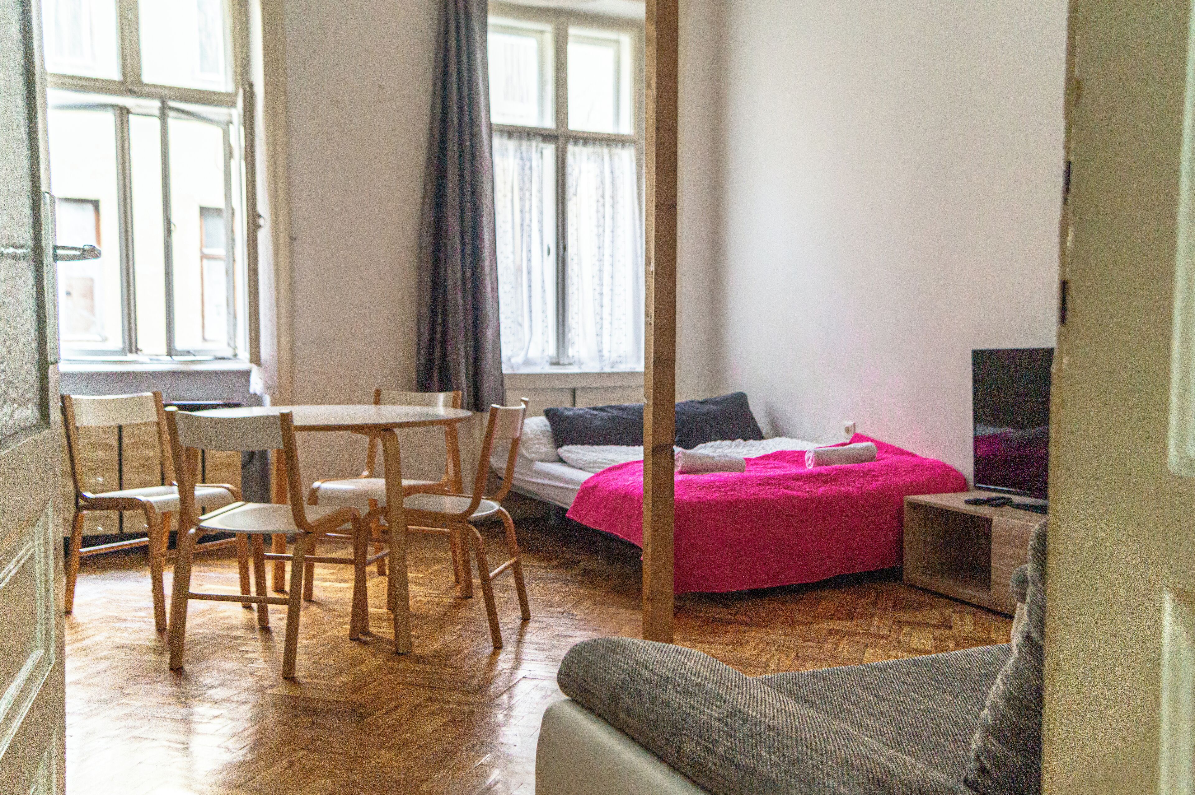 A room with a bed, a TV, a table with chairs, and a window with curtains.