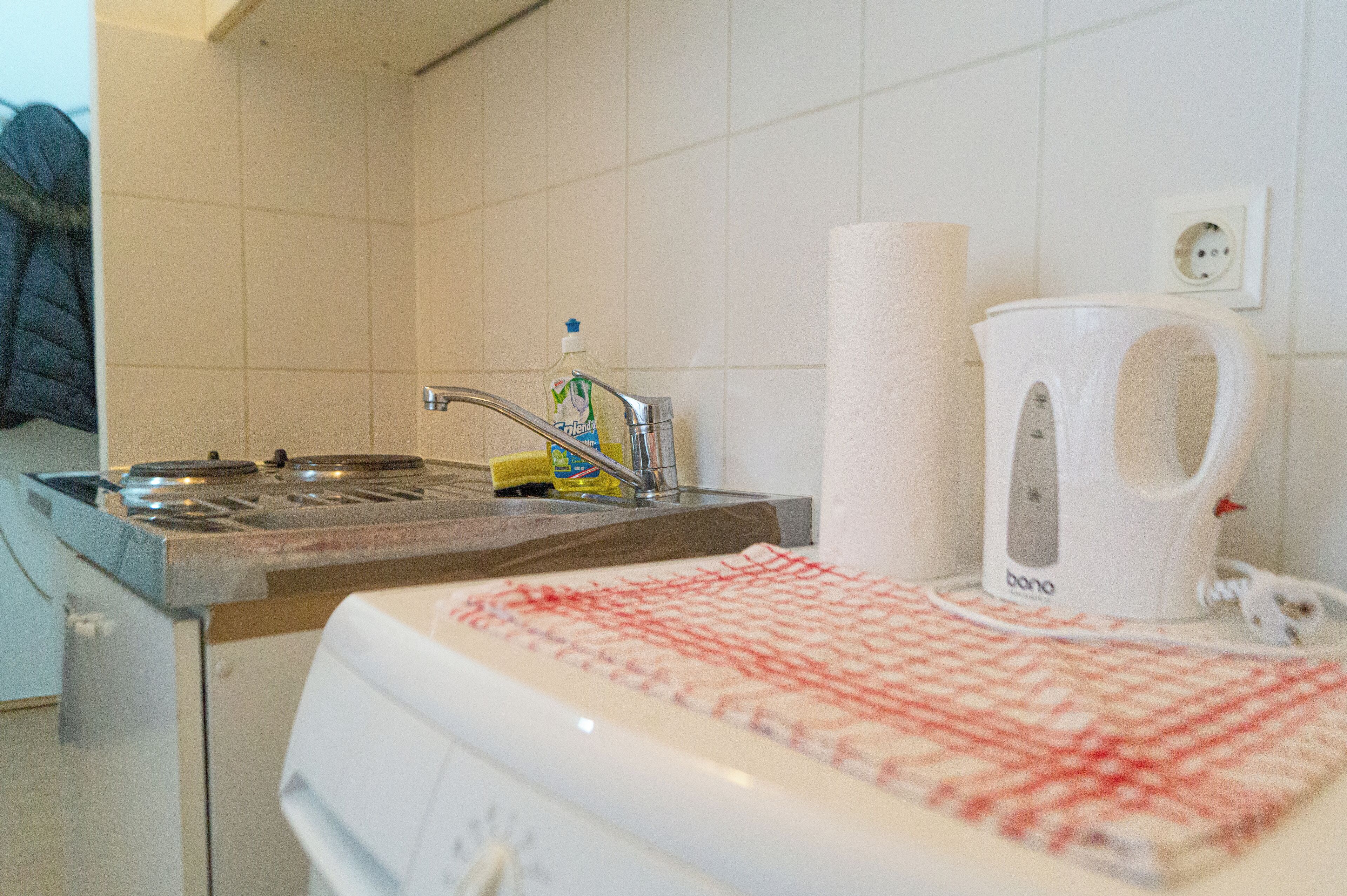 Una cocina con un lavabo, una botella de detergente, un dispensador de toallas de papel y una tetera blanca.