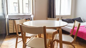 Quarto com uma mesa redonda e quatro cadeiras, uma cama com manta rosa e uma janela com cortinas.