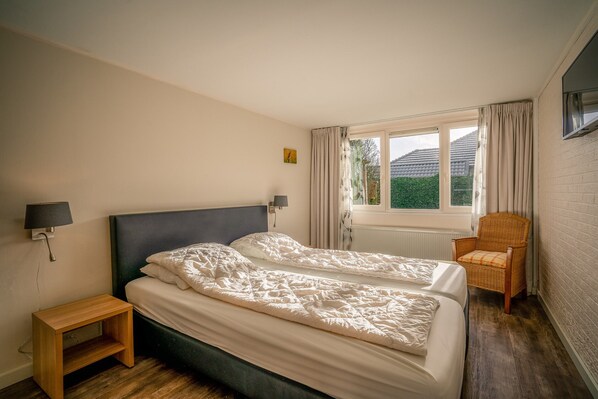 Una camera da letto con due letti, un comodino, una sedia, una finestra con tende e una TV a parete.