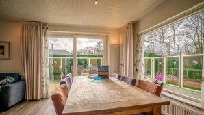 A dining area with a large wooden table, cushioned chairs, and a view of a garden through large windows. - Modernly Furnished 6-person Bungalow in the Seaside Resort of De Koog (De Koog)