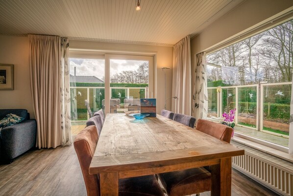 Un espace repas avec une grande table en bois, des chaises confortables et une vue sur un jardin grâce à de grandes fenêtres.