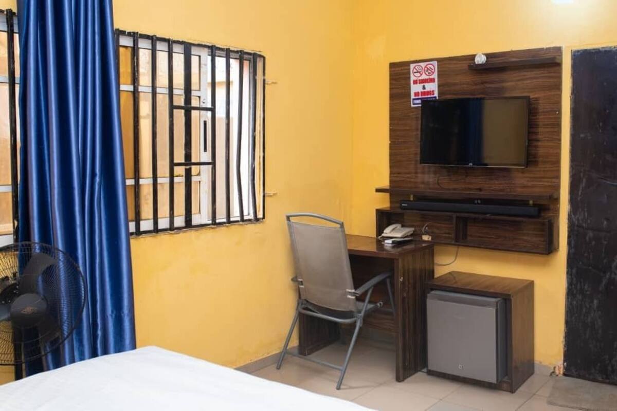 A small room with a bed, a desk with a chair, a TV mounted on the wall, a small refrigerator, and a window with bars.