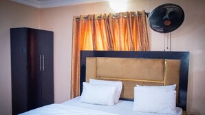 A hotel room with a bed, a wardrobe, a headboard, and a standing fan. - PROMISE HOTELS HOMES AND APARTMENT (Lagos)