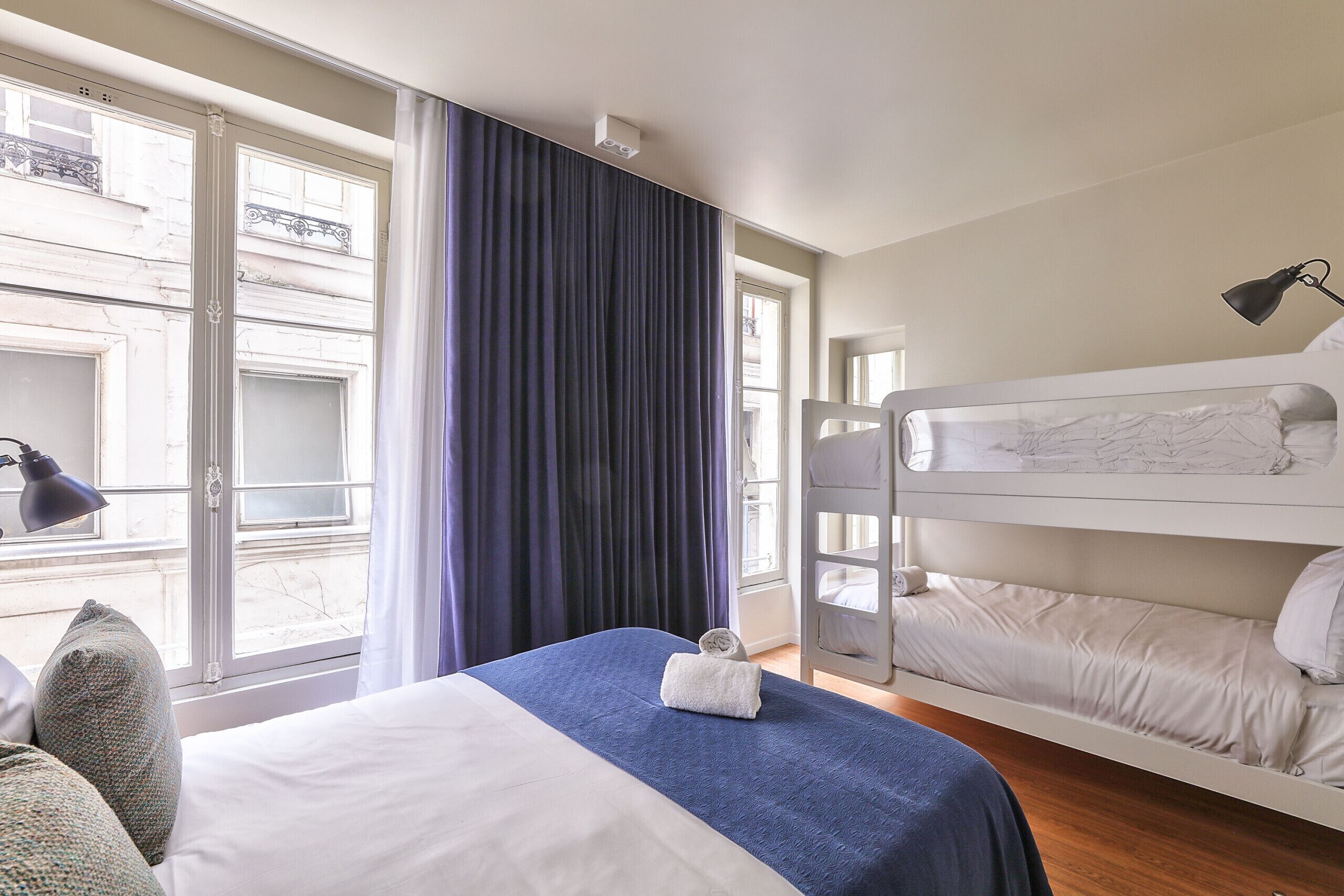 Een hotelkamer met een bed, een raam met gordijnen, een stapelbed en een bureaulamp.