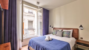 A hotel room with a large bed, a wooden nightstand, a window with curtains, and a view of a building outside.