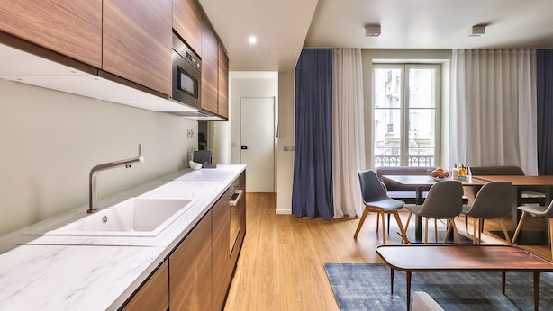 A modern kitchen with wooden cabinets, a stainless steel sink, and a built-in oven. There is a dining area with a table and chairs, and a view of the city through a window with curtains.