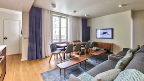 A modern living room with a sofa, a dining table, and chairs. There is a TV mounted on the wall and a window with curtains.