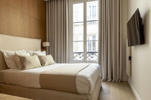 Habitación de hotel con cama, cortinas, televisor montado en la pared y ventana con vista a edificios.