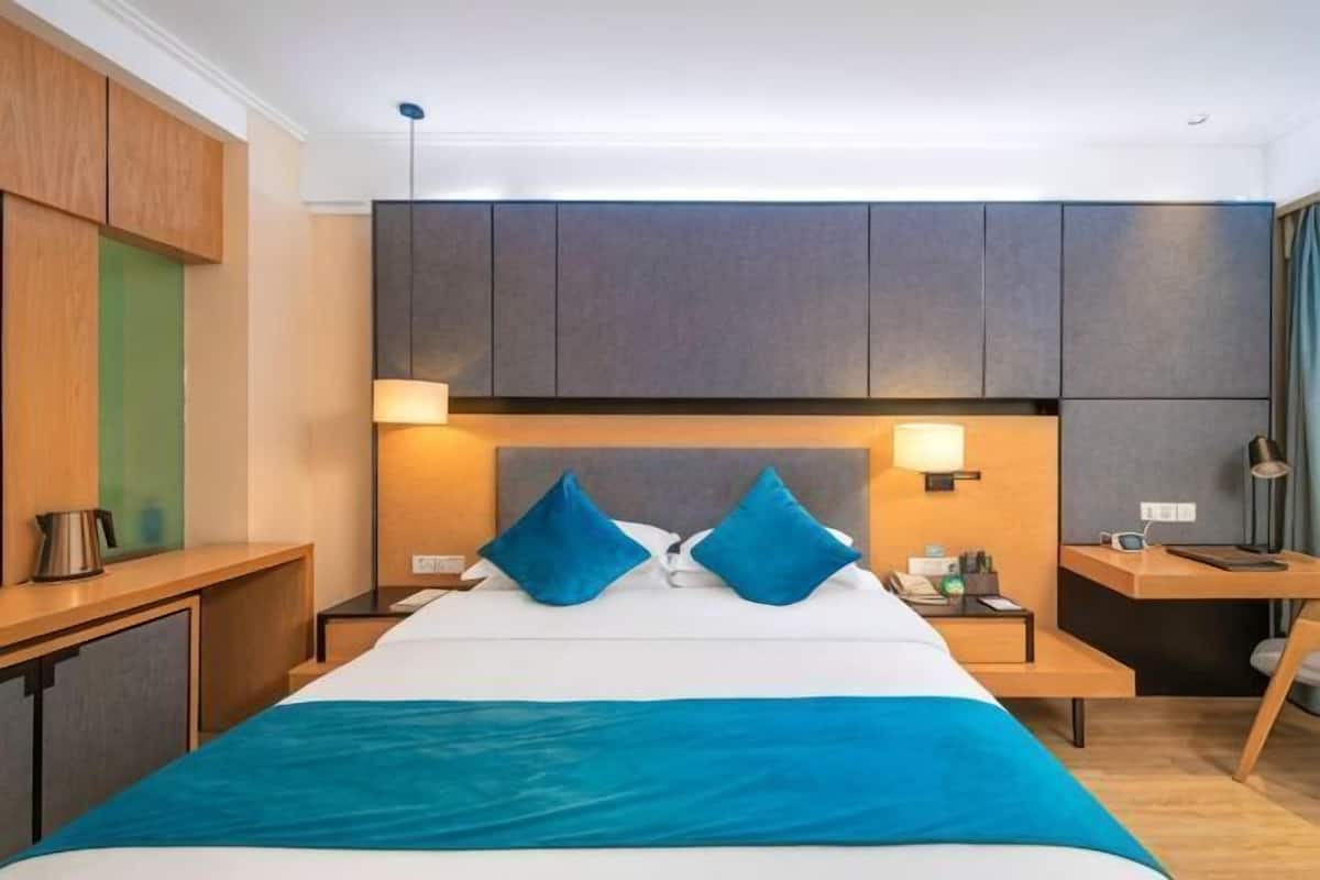 A modern hotel room with a large bed, bedside tables, a desk, and a headboard with built-in lighting.