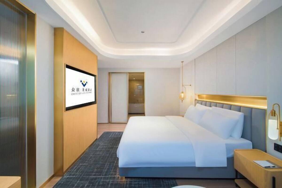 A modern hotel room with a large bed, a bedside table, a wall-mounted TV, and a door leading to another room.