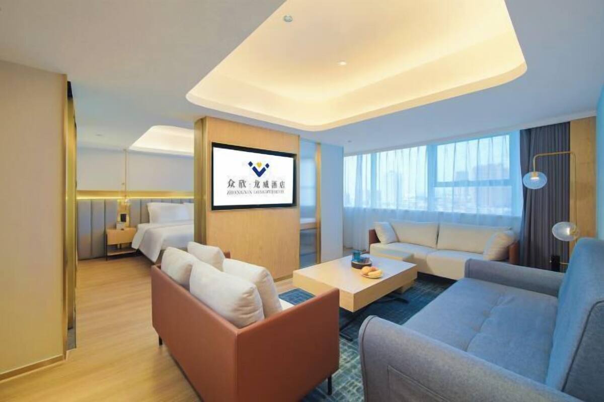 A modern hotel room with a sofa, a coffee table, a TV mounted on the wall, and a large window providing natural light.