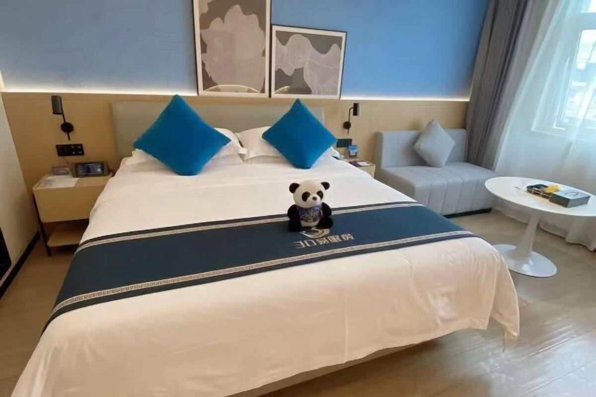 A hotel room with a bed, a nightstand, a sofa, a small round table, and a panda plush toy on the bed.