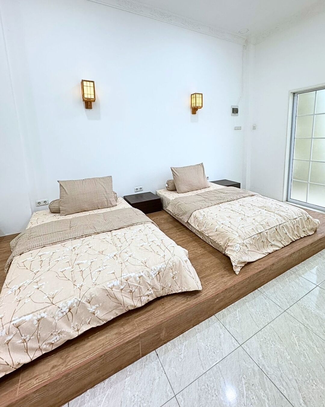 Una habitación de hotel con dos camas, cada una con una colcha de estampado floral y almohadas a juego. Hay lámparas de pared sobre cada cama, y la habitación tiene un gran ventanal corredera a la derecha.