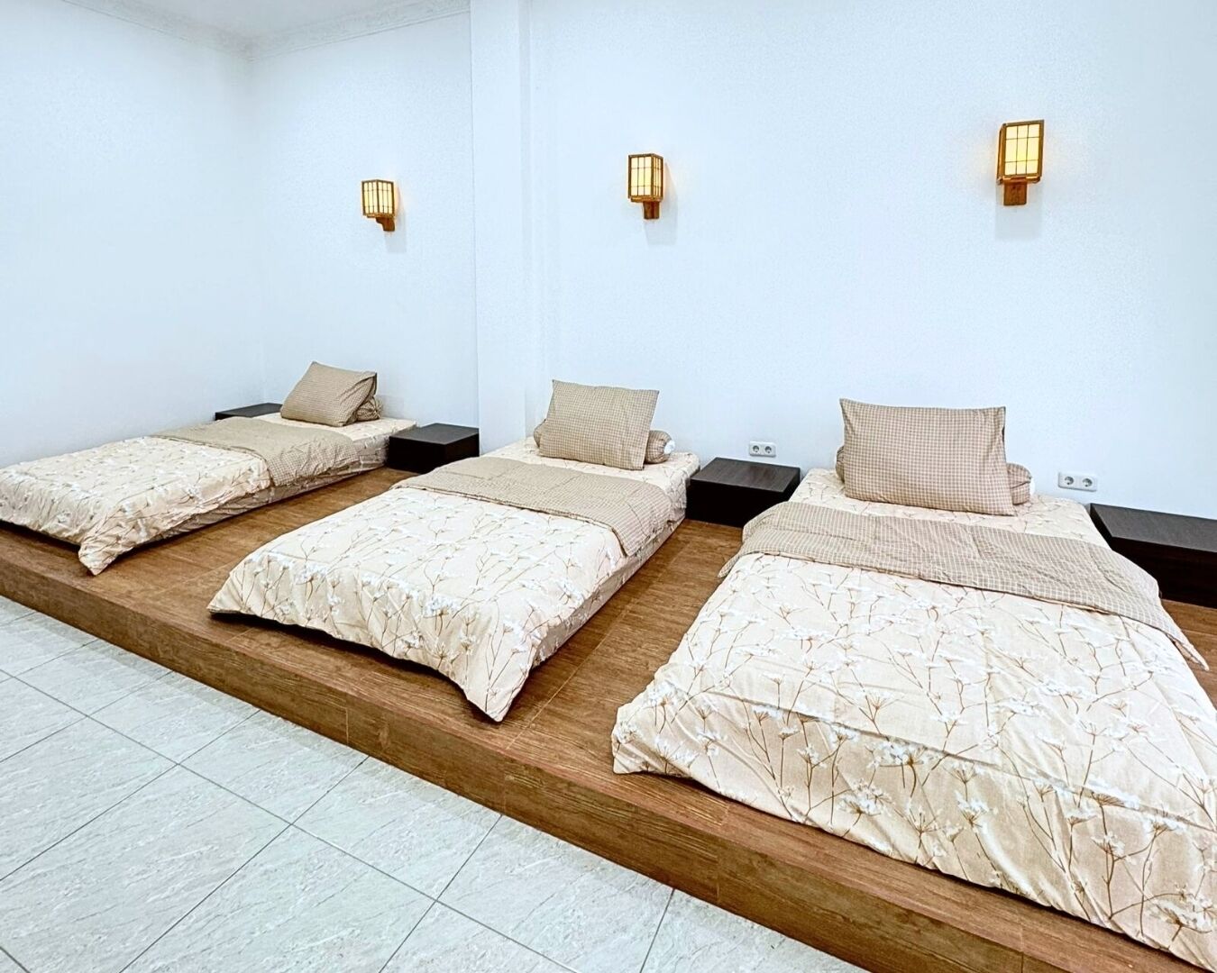 Een hotelkamer met drie eenpersoonsbedden, elk voorzien van een houten hoofdeinde, een nachtkastje en een wandlamp.