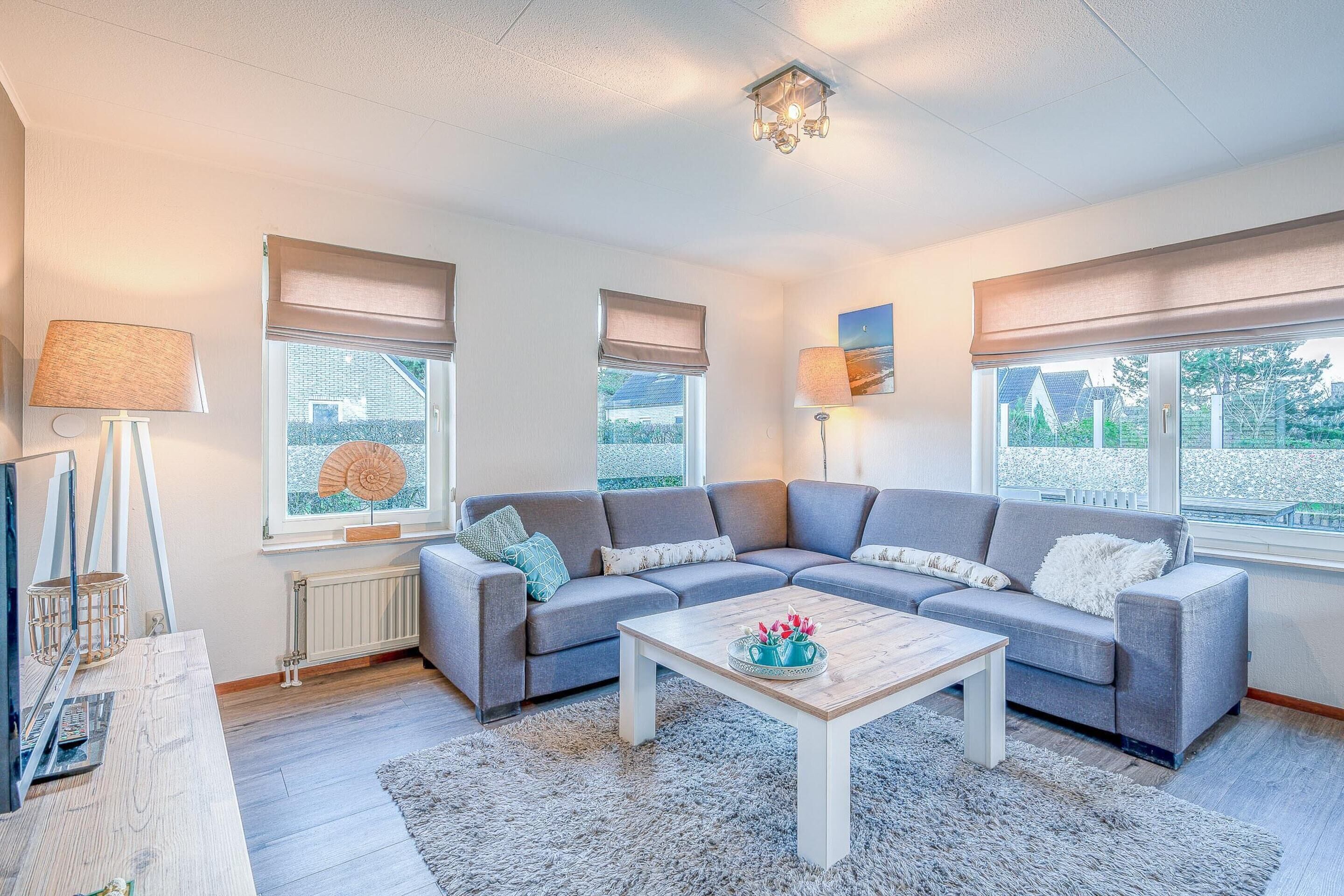 Uma sala de estar moderna com um sofá seccional cinzento, uma mesa de centro de madeira e uma janela grande com vista para vegetação.