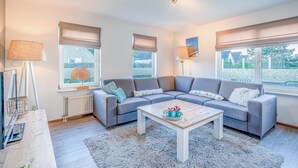 A modern living room with a grey sectional sofa, a wooden coffee table, and a large window with a view of greenery. - Charming Villa With Wood Stove Near the sea and Forest (De Koog)