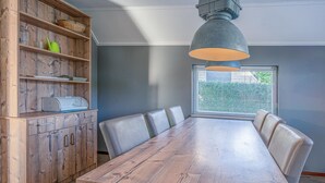 A dining area with a wooden table and chairs, a built-in wooden cabinet with shelves, and a window with blinds. - Charming Villa With Wood Stove Near the sea and Forest (De Koog)