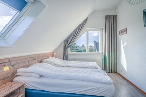 A bedroom with a sloped ceiling, a bed with white bedding, a wooden headboard, a bedside table with a lamp, and a window with curtains. - Charming Villa With Wood Stove Near the sea and Forest (De Koog)