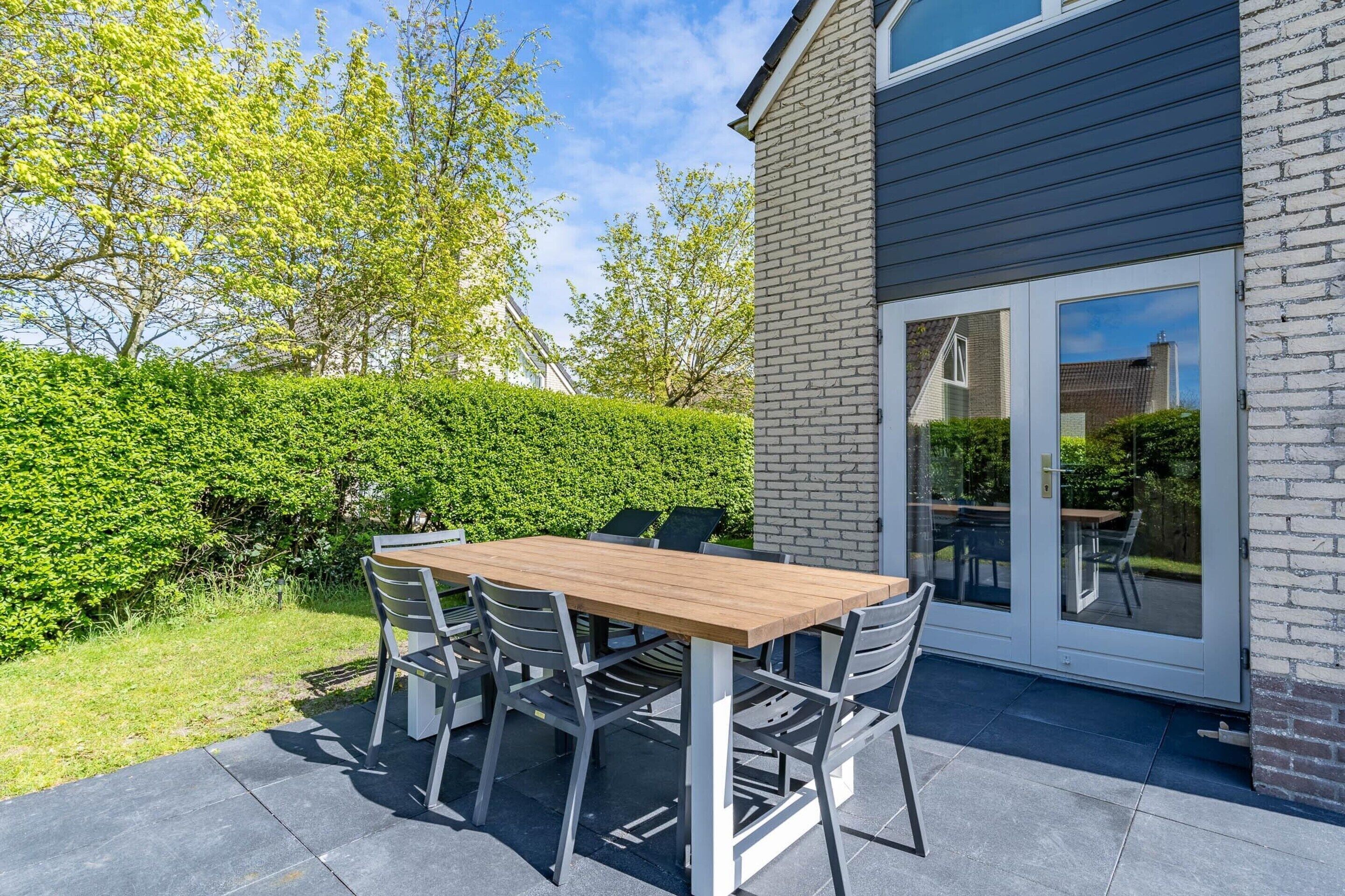 Une terrasse extérieure avec une table et des chaises en bois, un mur en briques et une porte vitrée donnant sur un espace abrité.